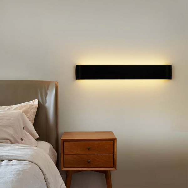 Rectangular black wall lamp, with integrated light. Hung on a white wall next to a bed, above a wooden nightstand.
