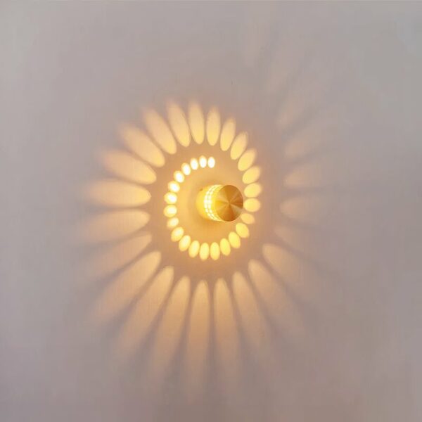 Wall light with effect, consisting of a grey metal cylinder, perforated with small holes, which lets the light spread in a spiral shape on the wall. On. Hung on a white wall.