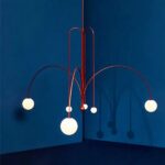Minimalist terracotta chandelier, composed of terracotta-red metal rods that form an original, modern structure. Two slender rods, one on the right, the other on the left, form an arc with white balls at the ends. The other slender rods also have white balls at the end. On a dark blue background.