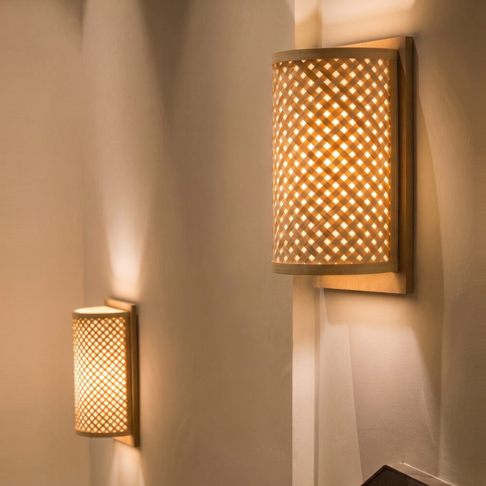 Two woven bamboo wall lights switched on against a light wall, mounted on a wooden back plate