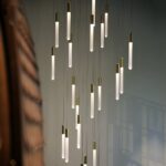 Close view of the staggered gold tube pendants against a grey-blue wall