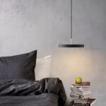 Black disc pendant with a brass stem glowing beside a bed with dark linen