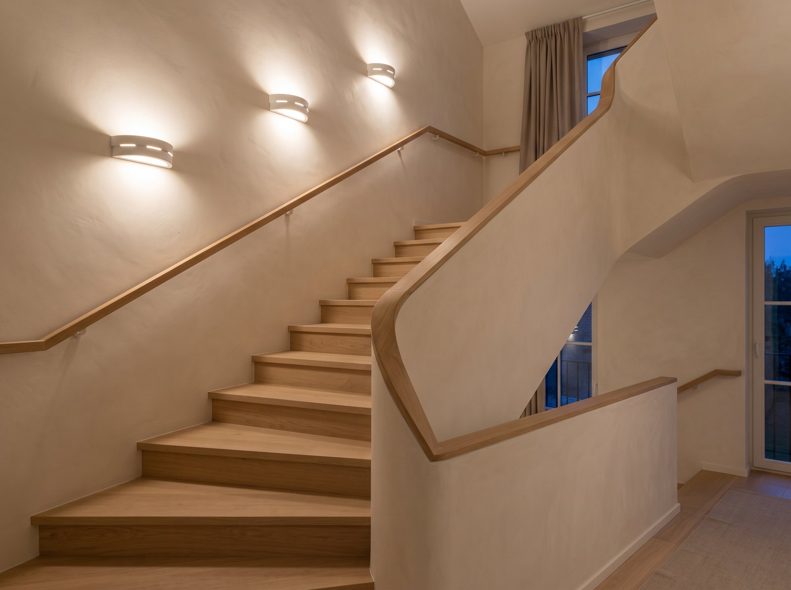 Quarter-turn oak staircase in the evening with three white ceramic half-moon wall lights lit along the slope, throwing light up and down a warm off-white wall