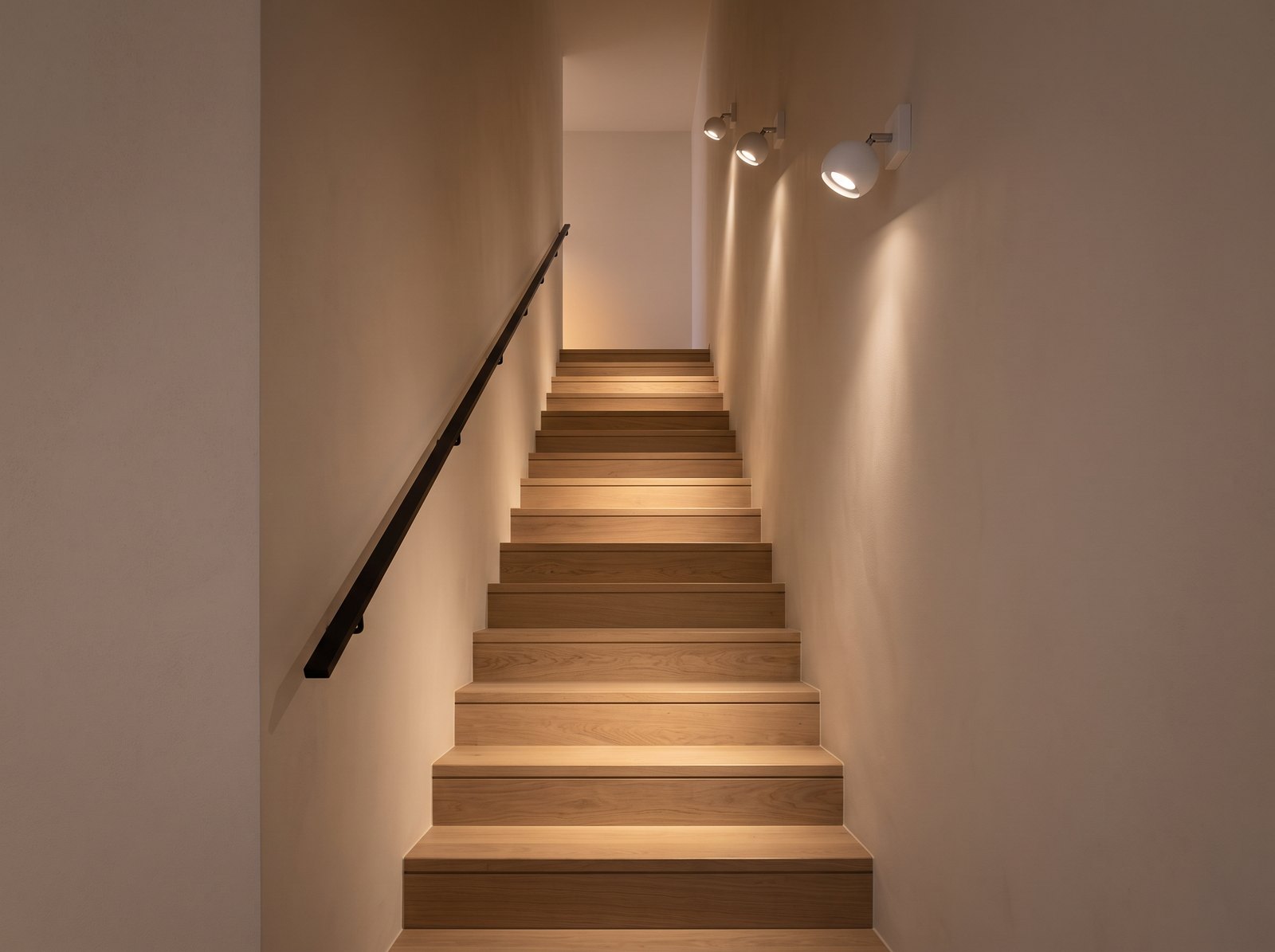 Narrow straight oak staircase between two pale walls, lit by three round white adjustable spot wall lights with their heads turned down towards the steps